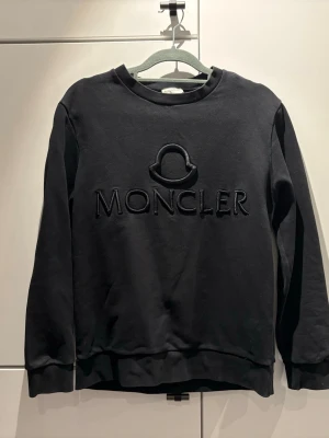 Svart Moncler sweatshirt med logga - Svart sweatshirt från Moncler med stor broderad logga och text på bröstet. Tröjan har rund halsringning, långa ärmar och ribbade muddar. Perfekt för en stilren och trendig look. Storlek M men passar även S jag är 174 och väger 64 den passar mig bra.