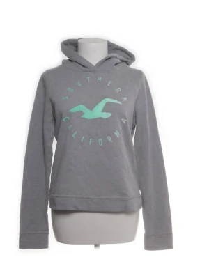 Grå hoodie från Hollister - Grå hoodie från Hollister med turkost tryck framtill där det står 'Southern California' och en stor måslogga. Hoodien har huva och långa ärmar. Perfekt för en avslappnad och cool stil. Mjuk bomullsblandning som känns skön mot huden. Inga defekter, förutom att märket har spruckit lite. Den passar också folk som har S.