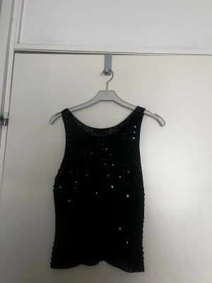 Svart paljettlinne från ellos - Snyggt svart linnetopp från ellos täckt med glittrande paljetter som ger en festlig look. Toppen är ärmlös med rund halsringning och har en figurnära passform. Perfekt för att sticka ut i mängden.