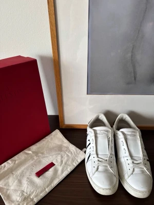 Vita Valentino Garavani sneakers - Snygga vita sneakers från Valentino Garavani med svart VLTN-logga på sidan. Skorna är i läder och har klassisk låg modell med vita snören och diskreta detaljer. Perfekta för dig som gillar stilrena och exklusiva sneakers. Allt og ingår