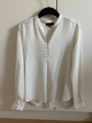 Vit blus med knappar från Selected Femme - Stilren vit blus från Selected Femme med lång ärm och dekorativa tygklädda knappar framtill. Blusen har en liten ståkrage och är tillverkad i ett mjukt, lätt material som ger ett elegant fall. Perfekt att matcha med jeans eller kjol.