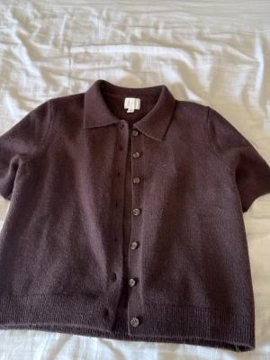 Brun kortärmad kofta från H&M - Mysig brun kofta från H&M med korta ärmar och knappar framtill. Koftan har klassisk krage och är stickad i ett mjukt material, perfekt för lager-på-lager. Enkel och stilren design som passar till många outfits.