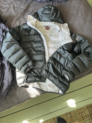 Grå pufferjacka från Colmar - Snygg pufferjacka från Colmar i grått med vit insida. Jackan har huva, dragkedja och elastiska muddar vid ärmslut och nederkant. Perfekt för kalla dagar och riktigt skön att bära. Klassisk quiltad design och logga på bröstet.