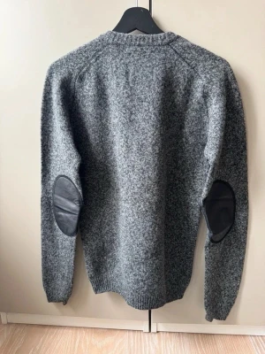 Acne cardigan - Gråmelerad V-ringad ulltröja från Acne studios med svarta mockalappar på armbågarna och diskret broderad logga på bröstet. Tröjan har ribbade muddar och klassisk passform, perfekt för en stilren look.