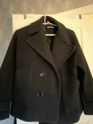 Svart kort kappa från Design by SI - Stilren svart kappa med breda slag, två stora knappar framtill och rymliga fickor. Klassisk modell med rak passform och siden tyg insida. Perfekt för kyliga dagar och enkel att matcha med olika outfits. Lite nopprig på utsidan vid vissa ställen. 