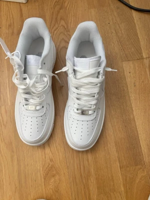 Nike Air Force 1 vita sneakers - Klassiska helvita Nike Air Force 1 sneakers med lågt skaft. Skorna har perforerad tå, bred sula och ikonisk Swoosh-logga på sidan. Tillverkade i läder med textilfoder och plös. Perfekta för en clean och stilren look.