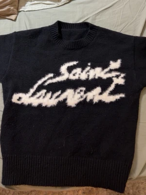 Svart Saint Laurent stickad tröja - Stickad svart tröja från Saint Laurent med vit, fluffig logotyp framtill i grafisk stil. Tröjan har rund halsringning och ribbstickade muddar. Perfekt för dig som gillar statement-plagg och exklusiv streetstyle.