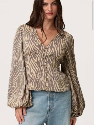 Zebramönstrad blus med puffärm - Trendig blus med zebramönster i beige och brunt. Djup V-ringning, knappar framtill och långa puffärmar ger en cool och modern look. Perfekt att styla med jeans eller kjol för en snygg outfit.