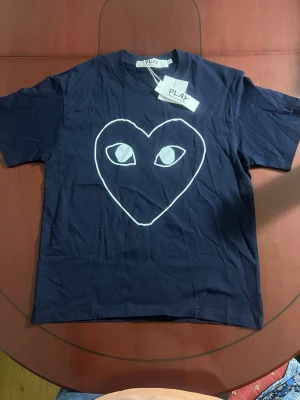 Mörkblå Comme des Garçons PLAY t-shirt - Säljer en mörkblå t-shirt från Comme des Garçons PLAY med det ikoniska hjärtmotivet med ögon på bröstet. T-shirten har korta ärmar, rund halsringning och är gjord i mjuk bomull. Perfekt för dig som gillar streetwear och vill sticka ut.