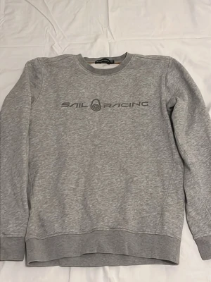 Grå sweatshirt från Sail Racing - Snygg grå sweatshirt från Sail Racing med rund halsringning och tryckt logga på bröstet. Tröjan har ribbade muddar vid ärmslut och nederkant, perfekt för en avslappnad och sportig stil.