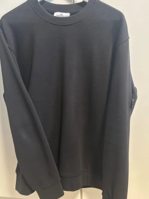 Svart sweatshirt från Stobe island blacked out - Säljer en svart sweatshirt från Stone Island med klassisk rund hals och ribbade muddar. Tröjan har den ikoniska Stone Island-loggan på vänster ärm. Perfekt för dig som gillar stilrena och exklusiva plagg.