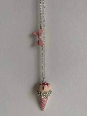 Halsband med glass-strut och rosett - Sött halsband med en silverfärgad kedja och ett hänge i form av en rosa glass-strut med vita och röda detaljer. På kedjan sitter även en rosa rosett. Perfekt accessoar för dig som gillar gulliga och lekfulla smycken.