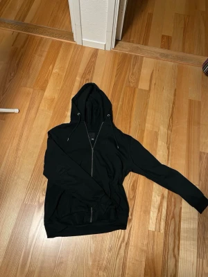 Svart hoodie med dragkedja från 157 - Svart hoodie från 157 i storlek M med dragkedja framtill och justerbar huva med snören. Mjuk insida och ribbade muddar vid ärmslut och nederkant. Perfekt för en chill och avslappnad stil.