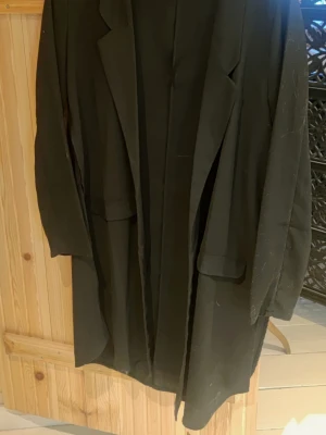 Svart lång kappa med klassisk krage - Stilren svart kappa i längre modell med klassisk krage och raka ärmar. Kappan har två fickor framtill och en enkel, tidlös design som passar till många olika outfits. Perfekt för dig som gillar minimalistisk och clean stil.