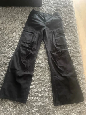 Svarta cargobyxor med vida ben - Säljer ett par svarta cargobyxor med bred passform och stora fickor på sidorna. Byxorna stängs med knapp och dragkedja. Perfekta för en avslappnad och trendig look. Materialet känns mjukt och följsamt.
