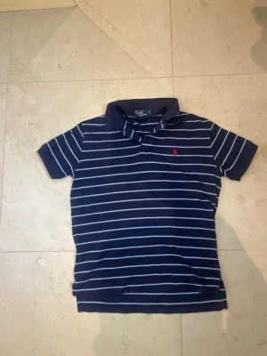 Marinblå randig pikétröja Ralph Lauren - Klassisk marinblå pikétröja från Ralph Lauren med vita horisontella ränder och röd broderad logga på bröstet. Tröjan har krage och korta ärmar, perfekt för en avslappnad men ändå stilren look.