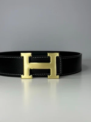 Svart bälte med guldfärgat H-spänne - Snyggt svart bälte med ett ikoniskt H-format guldfärgat spänne. Bältet är gjort i exklusivt skinn med tydliga sömmar längs kanterna och har en stilren, klassisk look som passar till det mesta.