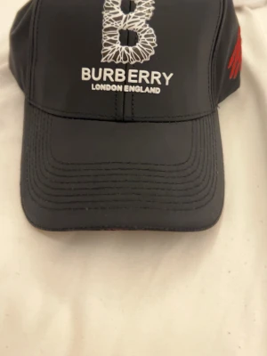 Svart keps från Burberry - Snygg svart keps från Burberry med vit broderad logga och texten 'BURBERRY LONDON ENGLAND' framtill. Justerbart spänne baktill och diskret röd detalj på sidan. Tillverkad i ett lätt och slitstarkt material som ger en modern look.