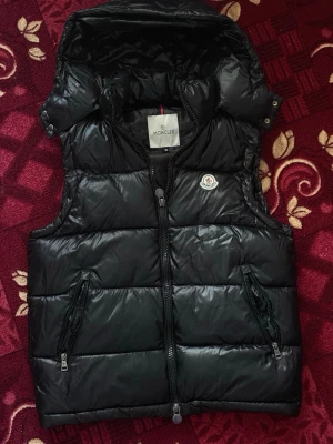 Moncler Vest Svart  - Säljer en svart, blank dunväst från Moncler i mycket bra skick. 100% äkta med NFC/QR-verifiering, originalmärke på bröstet och dragkedjor i toppskick. Varm men lätt och sitter riktigt snyggt – perfekt för vår, höst eller över hoodie på vintern. Ett exklusivt plagg som lyfter hela outfiten, först till kvarn!