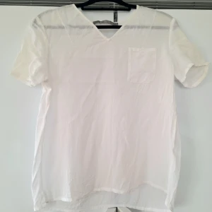 Vit kortärmad blus med V-ringning från Tiger  - Stilren vit blus med korta ärmar och.         V-ringning. Blusen har en bröstficka och är tillverkad i ett tunt, lätt material som ger en luftig känsla. Perfekt att matcha med jeans eller kjol för en fräsch look. Ber om ursäkt för ostruken. Den är samma vit hela men skuggorna gör att den ser lite gul ut på ärmarna. Det är den ej.