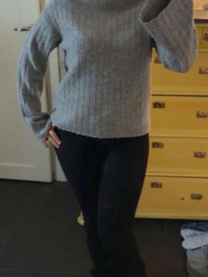 Grå ribbstickad polotröja - Mysig grå stickad ”polotröja” med ribbad struktur och långa ärmar. Tröjan har en avslappnad passform och hög krage som håller dig varm under kyliga dagar. Perfekt att matcha med jeans eller kjol för en stilren look.