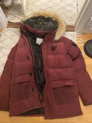 Vinröd dunjacka med fuskpäls - Snygg vinröd dunjacka från SikSilk med stor huva och fuskpälsdetalj. Jackan har två stora fickor framtill, svart innerfoder och broderad logga på bröstet. Perfekt för kalla vinterdagar och riktigt varm.
