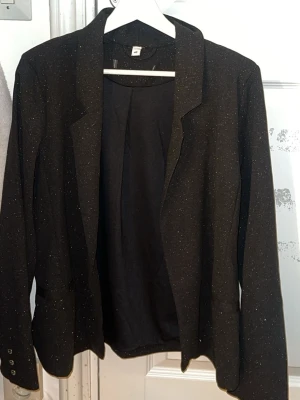 Svart glittrig kavaj med smal krage - Snygg svart kavaj med diskret glittrigt tyg och smal krage. Kavajen har långa ärmar med knappar vid ärmsluten och en klassisk passform. Perfekt för att lyfta en outfit med lite extra stil.