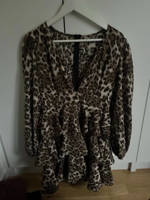 Leopardmönstrad volangklänning - Kort klänning med leopardmönster och flera volanger. Djup v-ringning och långa ärmar med lätt puff. Klänningen har dragkedja i ryggen och är tillverkad i ett glansigt, mjukt material som ger en lyxig känsla.