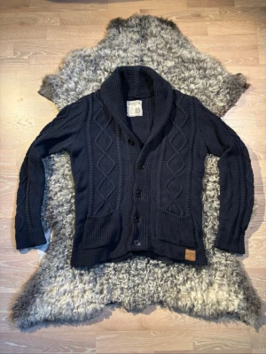 Mörkblå kabelstickad kofta - Snygg mörkblå kabelstickad kofta med sjalkrage och stora knappar framtill. Koftan har två framfickor och ribbade muddar. Tjockt garn och klassiskt mönster ger en tidlös look. Perfekt för lager på lager och chill stil.