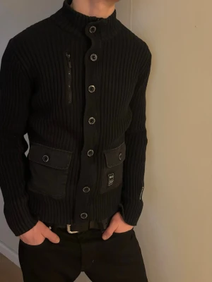 Svart ribbstickad kofta  - Snygg svart ribbstickad kofta med  krage, stora knappar framtill och två  ficko. Detaljerad med en bröstficka med dragkedja och diskret logga på ena fickan. Storlek m, sann i storlek, sitter såhär på 188cm. Mycket bra skick!