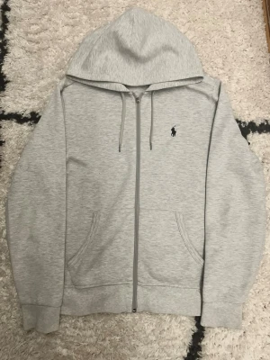 Grå zip hoodie från Polo Ralph Lauren - Defekt på 4 bilden Snygg ljusgrå hoodie med dragkedja från Polo Ralph Lauren. Klassisk design med två fickor fram, justerbar huva med snören och den ikoniska lilla loggan broderad på bröstet. Perfekt för en chill och stilren look.