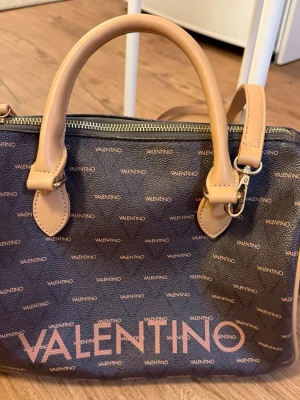 Brun axelväska från Valentino - Snygg brun axelväska från Valentino med beige handtag och detaljer. Väskan har ett mönster med små Valentino-loggor över hela ytan och en stor logga framtill. Tillverkad i konstläder med dragkedja upptill och avtagbar axelrem. Den har en liten repa på sidan. Nypris: 1495kr 