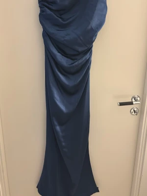 Marinblå balklänning i satin - Elegant marinblå balklänning i glansigt satinmaterial med draperad framsida. Klänningen har en figurnära passform och faller snyggt längs kroppen. Perfekt för speciella tillfällen när du vill känna dig extra fin.