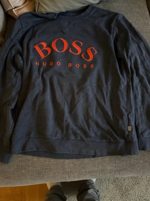 Mörkblå sweatshirt från Hugo Boss - Snygg mörkblå sweatshirt från Hugo Boss med stort rött BOSS-tryck på bröstet. Tröjan har rund halsringning och ribbade muddar vid ärmslut och nederkant. Perfekt för en avslappnad och trendig look.