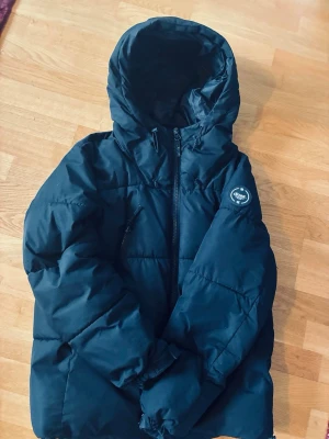 Svart pufferjacka med huva - Zara - Snygg svart pufferjacka från Zara med stor huva och dragkedja framtill. Jackan har en bröstficka med dragkedja och en rund logotyp på ärmen. Perfekt för kalla vinterdagar tack vare det tjocka, vadderade materialet. Innerfickorna behövs sys igen annars en grym jacka till bra pris. 