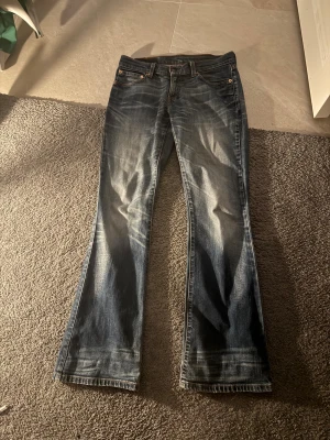 Blå bootcut jeans med slitningar - Säljer ett par blå bootcut jeans med snygga slitningar och ljusa tvättade partier. Klassisk femficksmodell med låg midja och dragkedja. Perfekta för en avslappnad och trendig look.