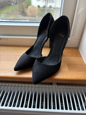 Svarta klackar från NLY - Säljer ett par svarta klackskor från NLY med spetsig tå och öppen sida. Skorna har en elegant siluett och är tillverkade i ett tygmaterial som ger en matt finish. Perfekta för dig som vill ha en stilren och trendig look.