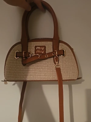 Beige och brun handväska  - Snygg handväska i beige flätat material med bruna läderdetaljer och guldfärgade metalldelar. Väskan har dubbla handtag, avtagbar axelrem och logotyp framtill. Perfekt för dig som gillar trendiga accessoarer.