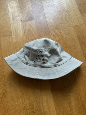 Beige bucket hat med metallöljetter från acne studio  - Snygg beige bucket hat i bomull med flera metallöljetter i ett triangulärt mönster på sidan för extra ventilation. Klassisk form med bred, mjuk brätte och avslappnad stil. Perfekt accessoar för soliga dagar och streetwear-vibe.
