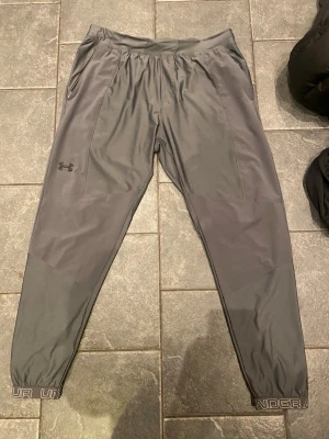 Gråa vindbyxor från Under Armour - Snygga grå mjukisbyxor från Under Armour med dragkedjefickor och logga på vänster lår. Byxorna har smal passform, resår i midjan och vid benslut samt detaljer med Under Armour-tryck längst ner. Perfekta för chill eller träning.