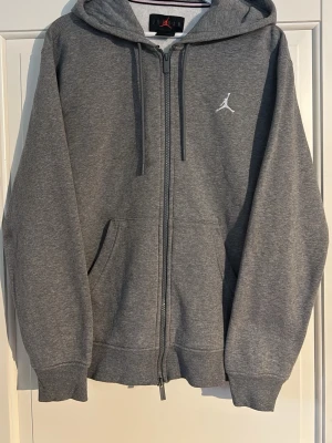 Grå zip hoodie från Jordan - Snygg grå hoodie från Jordan med dragkedja framtill och klassisk vit Jumpman-logga på bröstet. Hoodien har huva med dragsko och två fickor på sidorna. Tillverkad i mjukt bomullsmaterial som är perfekt för chill eller streetwear.liten fläck på armen men syn knappt