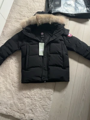  Canada Goose jacka - Snygg svart dunjacka från Canada Goose med avtagbar huva och äkta pälsdetalj. Jackan har klassisk patch på ärmen, dragkedja och knappar framtill samt två stora fickor. Perfekt för kalla vinterdagar och riktigt varm tack vare dunfyllningen.