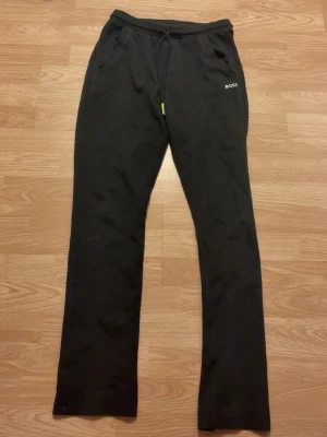 Svarta mjukisbyxor från BOSS - Säljer ett par svarta mjukisbyxor från BOSS med raka ben och resår i midjan med dragsko. Byxorna har fickor och en cool BOSS-logga i vitt och neongrönt på bakfickan. Perfekta för chill dagar eller när du vill ha en avslappnad stil.
