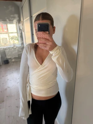Vit omlottopp med knytband - Snygg vit omlottopp med v-ringning och knytband i sidan. Långärmad och croppad modell som ger en fräsch och trendig look. Perfekt att matcha med jeans eller kjol för en cool stil.från lager 157