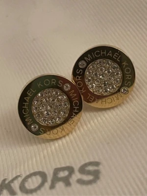 Guldtonade örhängen Michael Kors - Snygga runda örhängen från Michael Kors i guldtonad metall med gnistrande klara stenar i mitten och logotyp runt kanten. Perfekta för dig som gillar stilrena accessoarer med lite extra bling.