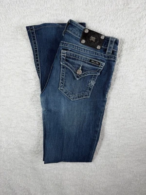 Mörkblå bootcut jeans från Miss Me - Snygga mörkblå bootcut jeans från Miss Me med dekorativa bakfickor och silverfärgade knappar. Jeansen har klassisk femficksdesign, kontrastsömmar och lätt slitning framtill. Perfekta för en trendig och avslappnad look.
