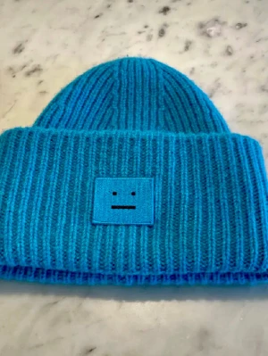 Ny Petrolblå  mössa Acne Studios  - Säljer en Ny blå ribbstickad mössa från Acne Studios med deras ikoniska ansiktslogga framtill, värmande material. Perfekt för dig som vill ha en stilren och trendig accessoar du aldrig tröttnar på. Pris i butik 1.700kr 