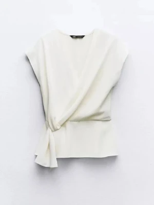 Vit omlottopp från Zara - Snygg vit omlottopp från Zara med knyt i sidan. Toppen har korta ärmar och en elegant v-ringning. Perfekt att styla med jeans eller kjol för en fräsch look. Tillverkad i ett mjukt material som känns skönt mot huden.