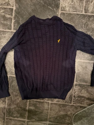 Mörkblå kabelstickad tröja  - Mörkblå kabelstickad tröja från pier one med rund halsringning och ribbade muddar. Tröjan har ett gult broderat logomärke på bröstet och är tillverkad i ett mjukt stickat material. Perfekt för en stilren och avslappnad look.