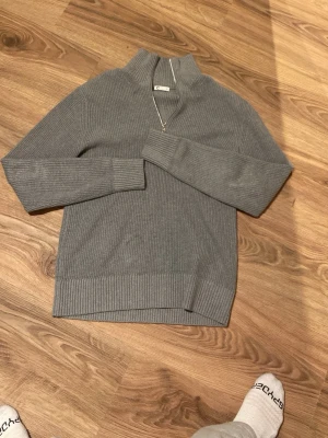 Grå ribbstickad tröja med halvzip - Snygg grå stickad tröja från C med ribbstickad struktur och halvzip i halsen. Tröjan har hög krage och långa ärmar, perfekt för lager på lager. 